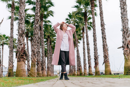 时尚的年轻女子在粉红色大衣摆在公园旁边的高大的绿色棕榈树照片