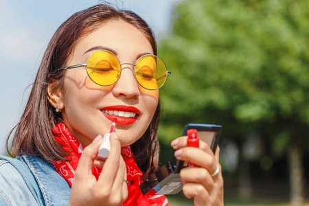 年轻的微笑的时髦的快乐的女人做化妆用口红和镜子户外