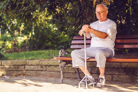 可怜的老人带着手杖坐在公园的长凳上