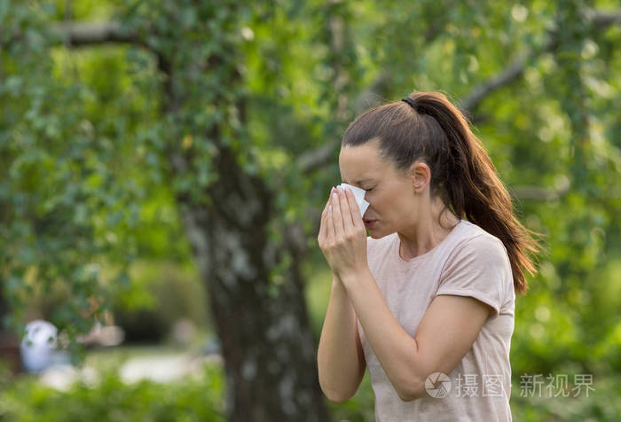 年轻漂亮的女人在公园里打喷嚏, 背景树.春季花粉过敏症状
