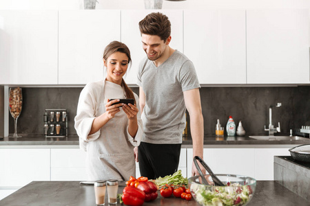 一对微笑的年轻夫妇在厨房一起做饭和使用手机的肖像