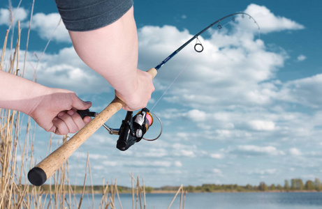 渔夫在纺纱时钓到一条鱼。手和线圈特写。弯曲的杆从咬。背景中的湖泊和多云的天空