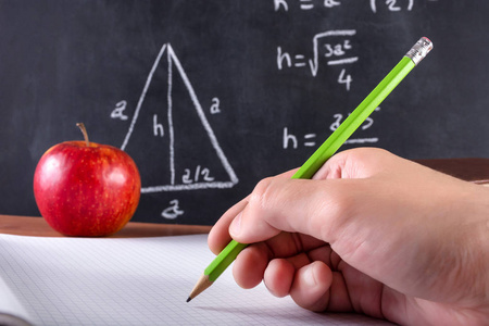 男学生手用绿木笔在笔记本上写字。红苹果和黑板的背景模糊不清。课堂教学理念。特写, 选择性聚焦