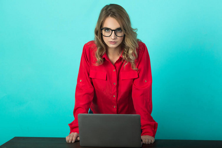 美丽的年轻女孩与眼镜和在红色的衬衫与笔记本电脑在蓝色背景