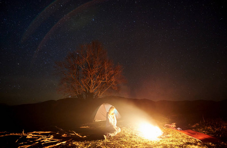 在山上露营的夜晚。女性游客在深暗的星空下休息, 周围有明亮的帐篷和大树的剪影。旅游和旅游概念