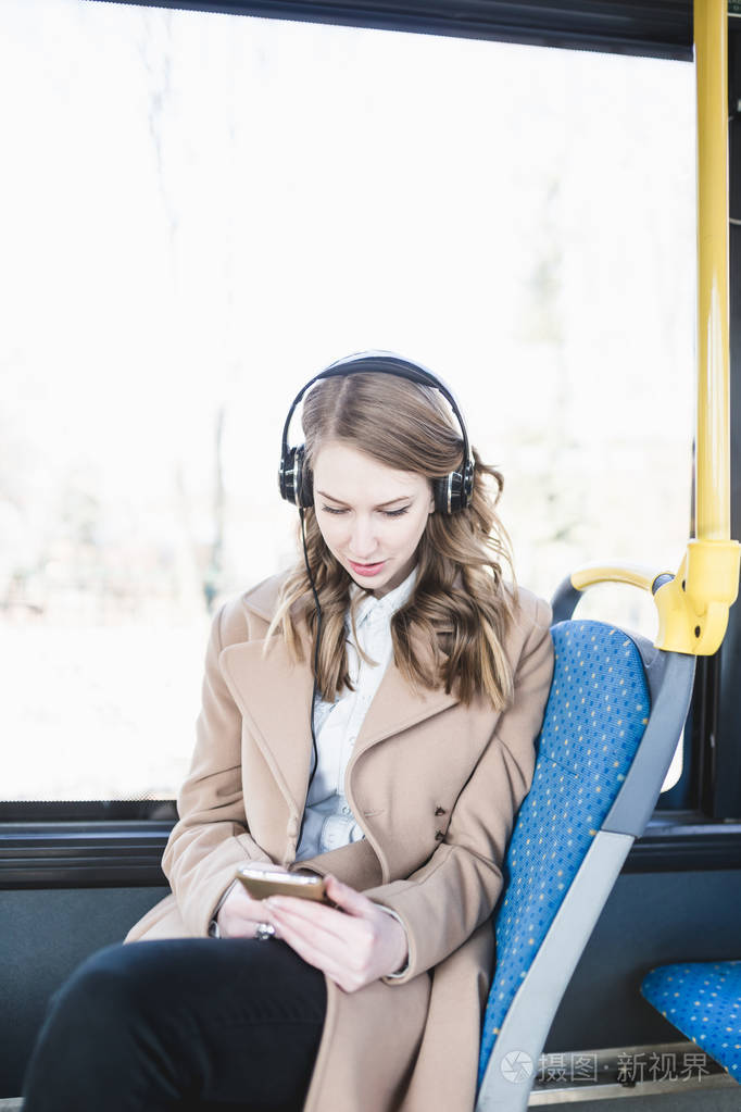 美丽快乐的年轻女子坐在城市公交车上, 看着手机和听音乐
