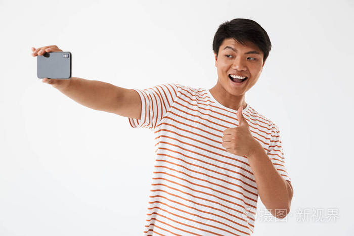 一个开朗的亚洲男子的肖像显示竖起大拇指的手势而采取自拍孤立的白色