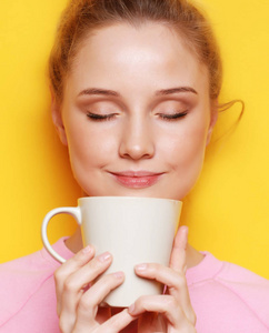 年轻的金发女人拿着白杯子喝茶或咖啡, 生活方式和食物的概念。黄色背景