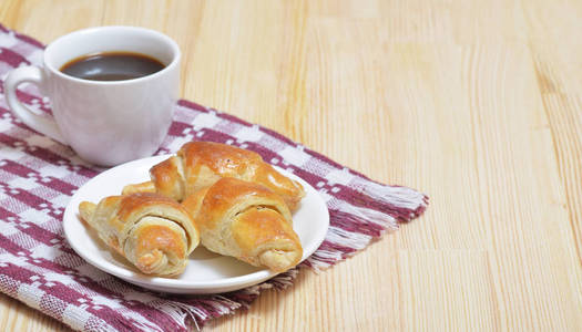 一小片羊角面包, 一杯咖啡, 在一张轻木桌上放着餐巾。