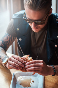 一个饥饿的时髦的家伙, 一个时髦的发型和胡子坐在桌子上, 决定在路边咖啡馆吃饭, 吃汉堡包