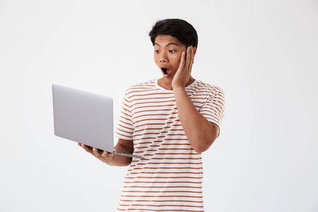 一个震惊的年轻亚裔男子的肖像看着笔记本电脑, 而站在白色隔离
