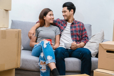 亚洲年轻夫妇坐在沙发上, 喝着放松后, 成功包装大纸板箱移动在新房子, 移动和房子狩猎的概念