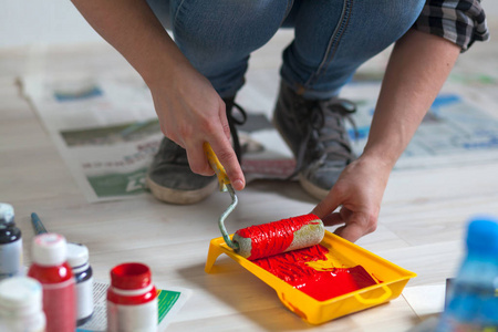 一个穿牛仔裤和运动鞋的女人在一个塑料托盘里拿起红色的油漆。画家准备粉刷墙壁修理孩子的房间。