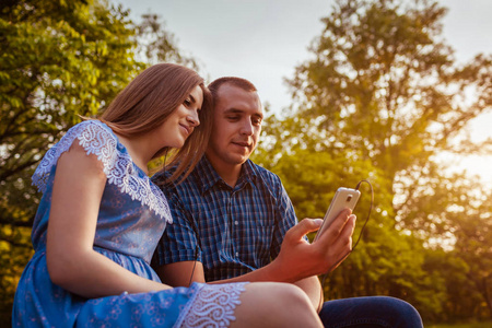 年轻夫妇听音乐通过耳机上的智能手机在春季公园日落。快乐的女人和男人拥抱