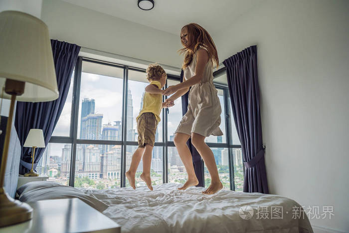 快乐的母亲和儿子跳上床的背景下的大窗口俯瞰摩天大楼和大城市