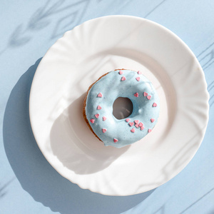 甜甜圈覆盖着蓝色的釉面, 在蓝色背景的白色盘子上洒上粉红色的小心, 表面上有图案的阴影。