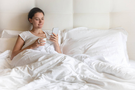 肖像漂亮的年轻女孩与智能手机在床上的早晨