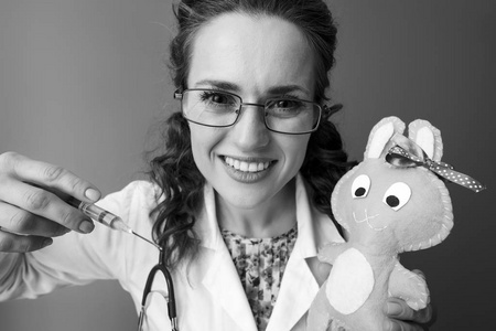 微笑的儿科医生妇女在白色的医疗长袍持有玩具和使用注射器反对背景