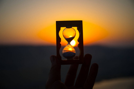 生命时间流逝的概念.手持沙漏与日落天空背景.选择性聚焦照片