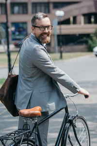 微笑英俊的生意人站立与自行车和看在街道的照相机在城市