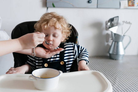 关心母亲喂她的小卷曲的儿子用有用的可口的汤, 家庭晚餐
