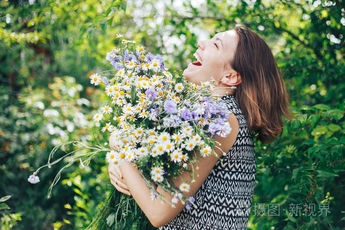 年轻美丽的女孩捧着野花的大花束甘菊看着相机夏天的时候绿草蓝花笑