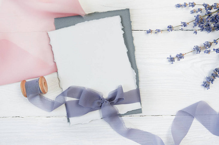 白色空白卡片丝带弓在粉红色和蓝色织品的背景与薰衣草花在白色背景。带信封和空白卡的样机。平躺。顶部视图