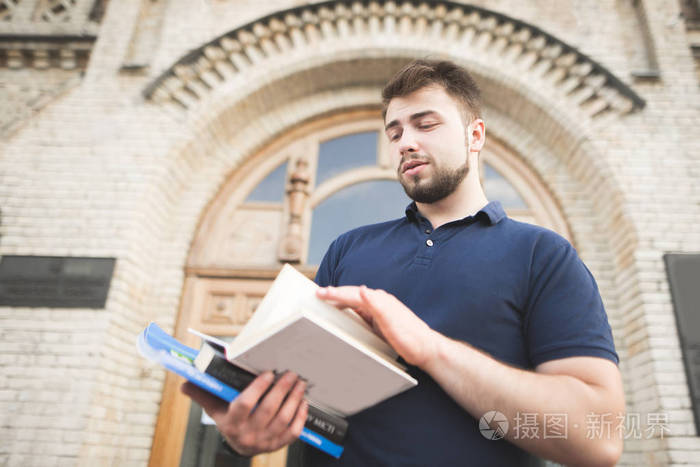 学生站在大学大楼前读这本书。一个人的肖像与胡子和书籍在手对旧建筑的背景