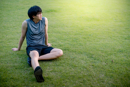 年轻的亚裔男子跑步后坐在草地上放松。户外运动理念下的健康生活方式