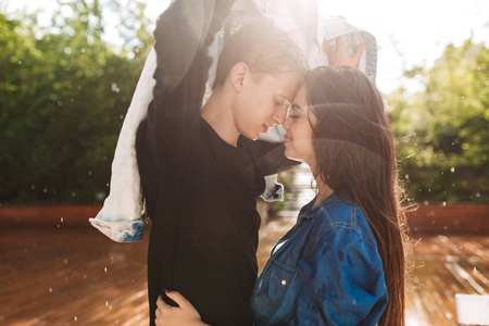 年轻的微笑夫妇的肖像覆盖自己的牛仔夹克, 而站在雨中公园。美丽的情侣一起共度时光