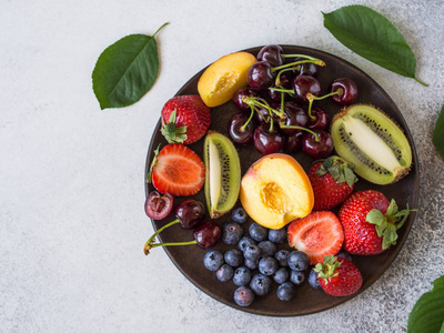 在灰色背景上的木盘子上的各种浆果和水果。草莓, 樱桃, 猕猴桃, 桃子, 蓝莓在盘子里。顶部视图