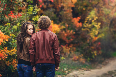 卷曲的头发的人转过身来, 女孩从他的肩膀后面窥视。可爱的情侣在美丽多彩的秋天树背景下