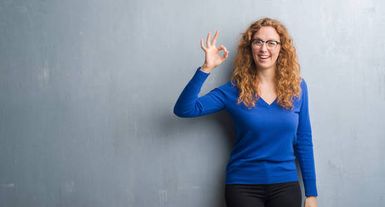 灰色垃圾墙上的年轻红头发的女人微笑积极做 ok 标志用手和手指。成功表达式