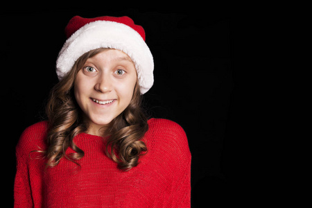 兴奋的小女孩穿着红毛衣和圣诞老人的帽子看着观众和笑。黑隔离。复制空间