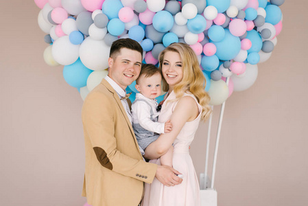 美丽的年轻父母微笑与他们的1岁的孩子在粉红色和蓝色气球背景。家庭外观。生日快乐派对