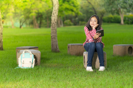 在阳光明媚的夏日, 学校公园里有一个数码平板电脑的亚洲学生女孩