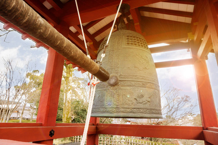 大黄铜佛教钟和日本寺庙的敲钟木材在明亮的蓝色天空与太阳和透镜光晕背景的红色亭子