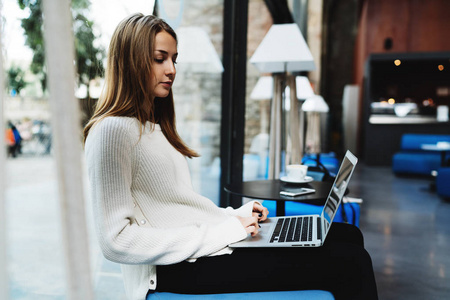 年轻的金发女郎坐在膝上膝上的笔记本电脑在一个大窗户内部的现代咖啡店