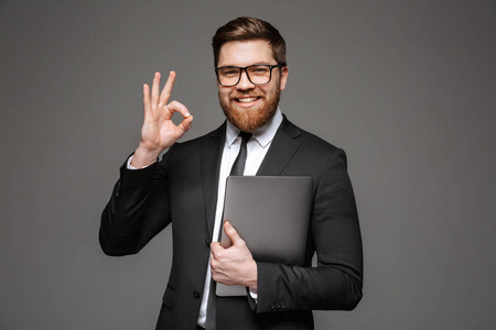 身着西装的微笑年轻商人的肖像手持笔记本电脑, 并显示在灰色背景下的 ok 手势隔离