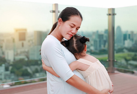 亚洲儿童女孩戴着皇冠拥抱她怀孕的母亲在屋顶上的建筑物日落