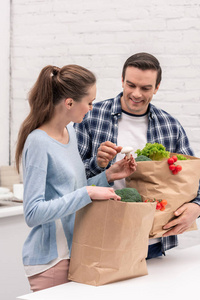 微笑的成人夫妇与纸袋子从杂货店在厨房里
