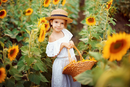 穿着白色连衣裙的小女孩, 一顶满是向日葵的草帽, 在一张向日葵的田野里微笑着看着照相机。