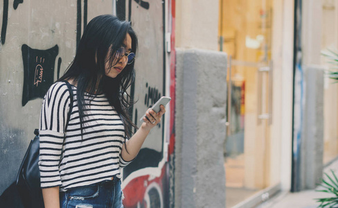 美丽的亚洲女性穿着眼镜和休闲服装站在街头背景与手机在她的手