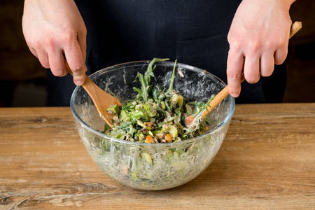 特写照片的男性手混合美味的绿色沙拉与芝麻菜和菠菜在玻璃碗与木勺和叉子在桌子上
