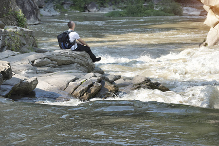 带着背包的年轻人坐在一条暴风雨的河岸上。阳光明媚的夏日