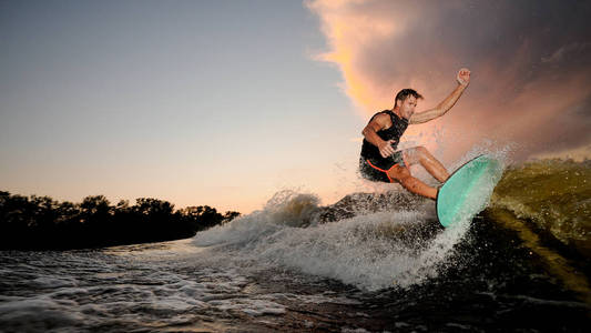 英俊的肌肉年轻男子 wakesurfing 下河波浪的背景下美丽的多云的天空在傍晚