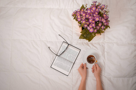 妇女的胳膊与杯子红茶, 杯子, 书和紫色的花在白色床上