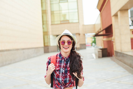 快乐的女性游客在太阳镜和帽子与背包走在街上的背景下, 建筑玻璃幕墙