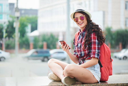美丽的微笑的女人在夏天帽子和太阳镜手持智能手机, 看着相机, 而坐在栏杆上