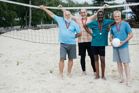 微笑的跨种族的老排球运动员以奖牌站立在沙滩上